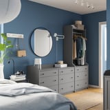 Moderní modrá ložnice s šedými zásuvkami a šatní skříní HAUGA, kulatým zrcadlem, bílou lampou, rostlinami a sluchátky na knihách.