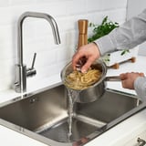 Osoba vypouštějící těstoviny v kuchyňském dřezu s moderní stříbrnou baterií.