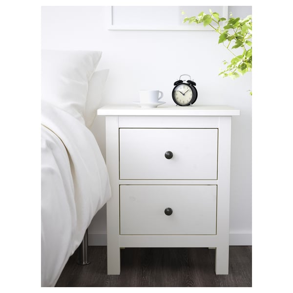 Noční stolek HEMNES s hodinami, zásuvkami, vedle postele s rostlinou.