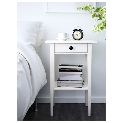 Bílý noční stolek HEMNES s černými hodinami, hrnkem na kávu, knihami. Vysoký, štíhlý, dřevěný design. Pohodlně se vejde vedle velké postele.