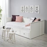 Bílá pohovka HEMNES s úložným prostorem, polštáři a přikrývkou. Malé odkládací stolky s hrnkem a knihou. Potisky motýlů na stěnách.