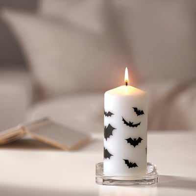 Bílá halloweenská svíčka HÖSTAGILLE, zdobená černými netopýry, je umístěna v průhledném skleněném držáku. Je malý, umístěný vedle zápalek na stole.