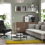 Moderní obývací pokoj s šedou pohovkou, barevným kobercem, skleněným stolem, dřevěnou skříní, koženým křeslem, policemi s knihami a světle zelenými stěnami.