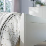 Útulná ložnice se světle modrou postelí IDANÄS. Květinové povlečení, bílá deka, plyšové čelo postele. Měkké přirozené světlo.