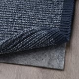 Tmavě modrý texturovaný koberec složený na světlé dřevěné podlaze, pod ním šedá protiskluzová podložka.