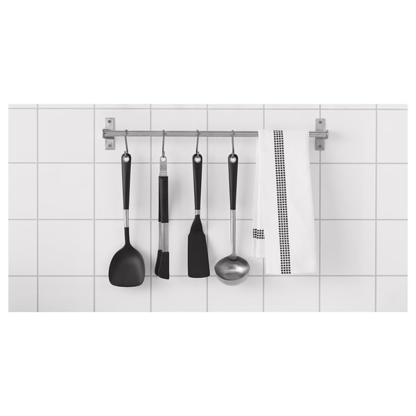 Černé nádobí IKEA na stříbrné kolejnici proti bílým dlaždicím - kuchyňské náčiní.