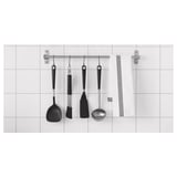 Černé nádobí IKEA na stříbrné kolejnici proti bílým dlaždicím - kuchyňské náčiní.
