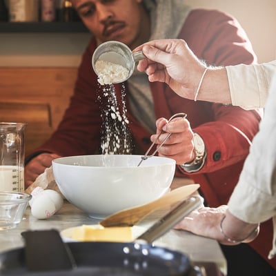 Člověk nasype mouku do mísy pomocí odměrky. Na stole je mísa, ingredience a kuchyňské náčiní.