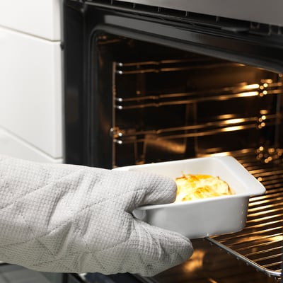 Ruka držící malou bílou porcelánovou misku s jídlem do trouby. Mísa má rukojeti a je součástí série IKEA 365+ pro bezpečné a snadné servírování.