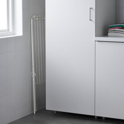 Bílý sušák, štíhlý, skládací vedle skříňky. Vysoký, vnitřní/venkovní, kompaktní, když se nepoužívá.