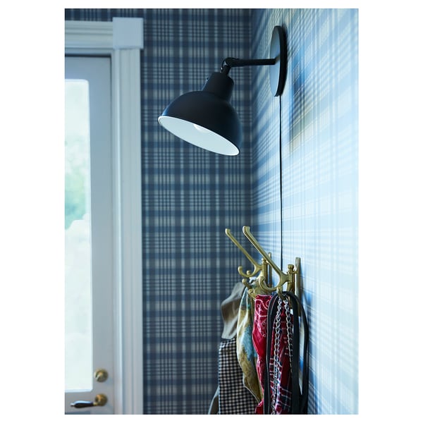 Černá nástěnná lampa nad zlatými háčky se zavěšenými předměty. Kostkovaná modrá stěna, bílé dveře.