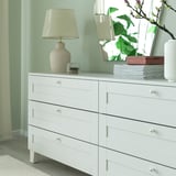 Bílá komoda se šesti zásuvkami. Dekorativní lampa, zrcadlo, hromada knih a váza. Knoflíky jsou bílé porcelánové.