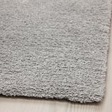 Měkký, plyšový, světle šedý koberec s mírným leskem, položený na dřevěné podlaze. Silný, útulný, recyklovaný polyester.