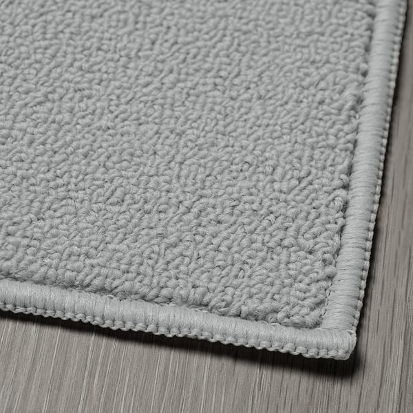 Detail texturovaného, šedého tkaného koberce se silným, robustním okrajem, položeného naplocho na dřevěné podlaze.