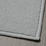 Detail texturovaného, šedého tkaného koberce se silným, robustním okrajem, položeného naplocho na dřevěné podlaze.