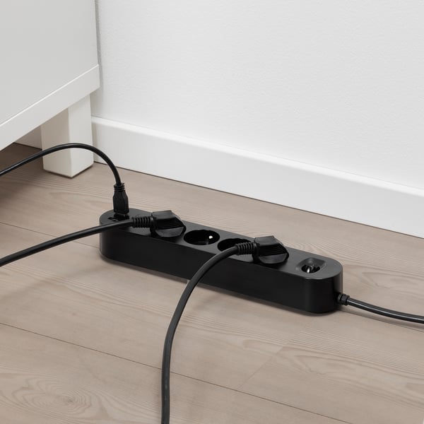 Černá multizásuvka s usb porty na dřevěné podlaze, zapojuje se do zdi, distribuuje energii do více zařízení.