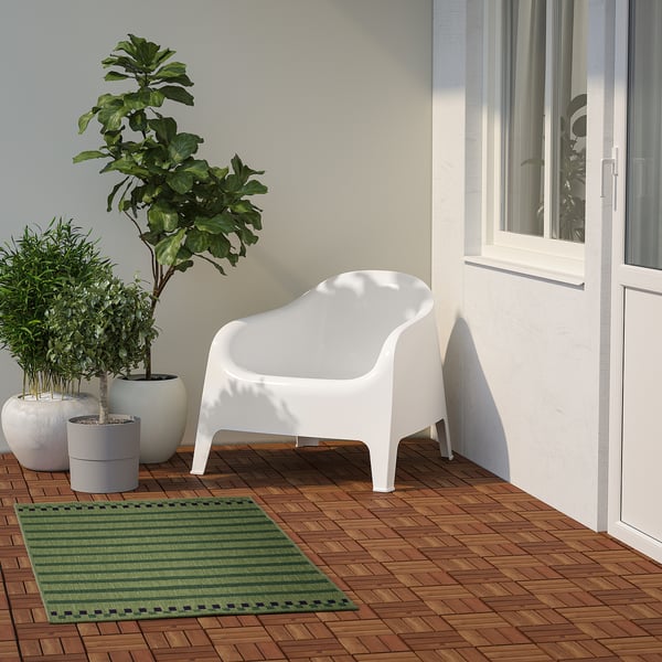 Malý zelený koberec KORSNING pod bílou židlí, obklopený rostlinami.