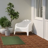 Malý zelený koberec KORSNING pod bílou židlí, obklopený rostlinami.