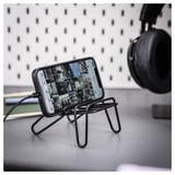 Černý stojan na mobilní telefon držící smartphone na stole, se sluchátky v pozadí.