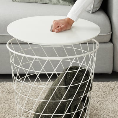 Osoba zvedající bílý stůl KVISTBRO s košíkem, který ukazuje jeho lehký a snadno přemístitelný design.