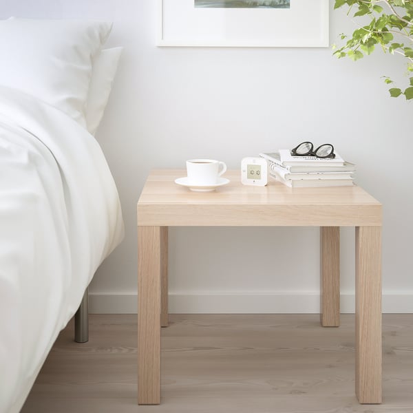 Odkládací stolek LACK jako noční stolek vedle postele, držící hrnek, knihy, brýle, doplňující bílé povlečení v minimalistické ložnici.