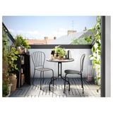 Malý balkon s černým kovovým stolem a dvěma židlemi, obklopený rostlinami v květináčích. Útulné městské venkovní prostředí.