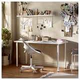 Světlý, bílý psací stůl LAGKAPTEN s minimalistickým skandinávským designem, s deskou z dřevovláknité desky a kovovými nohami, obklopený dekoracemi a uměním.