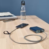 Dřevěný stůl s nabíjením telefonu přes modrý kabel LILLHULT z powerbanky, láhve na vodu a papírů v blízkosti.