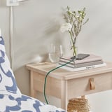 Noční stolek s telefonem nabíjeným přes zelený usb kabel, sklenice vody, knihy a váza s bílými květinami.