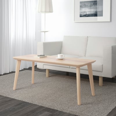 Minimalistický obývací pokoj s dřevěným konferenčním stolkem, hrnkem, poznámkovým blokem. Šedá pohovka, lampa, pruhovaný koberec.