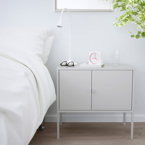 Bílá úložná skříňka IKEA LIXHULT vedle postele. Je kompaktní, má dvě dvířka a unese lampu, hodiny, brýle a vázu.