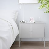 Bílá úložná skříňka IKEA LIXHULT vedle postele. Je kompaktní, má dvě dvířka a unese lampu, hodiny, brýle a vázu.