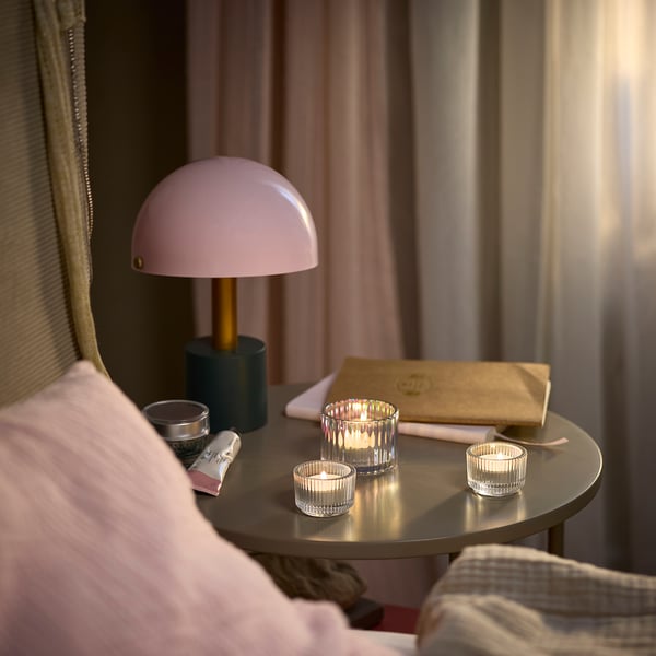 Čajové svíčky, lampa, kniha na malém stolku vedle růžové polštářové židle.