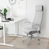 Moderní kancelářské uspořádání s ergonomickou židlí MARKUS z šedé síťoviny s vysokým opěradlem, bílým stolem, lampou a rostlinou. Židle je vysoká a poskytuje dobrou oporu zad.