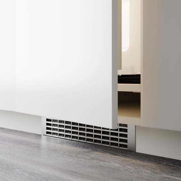 Malý větrací otvor instalovaný na základně bílé skříňky, pravděpodobně pro ventilaci nebo vedení kabelů v moderní kuchyni.