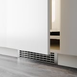 Malý větrací otvor instalovaný na základně bílé skříňky, pravděpodobně pro ventilaci nebo vedení kabelů v moderní kuchyni.