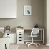 Minimalistická domácí kancelář se psacím stolem MITTCIRKEL ze světlého dřeva, bílou židlí a lampou. Béžové stěny s abstraktním uměním. Úhledné a organizované.