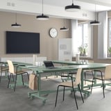 Moderní kancelář se zelenými skládacími stoly, židlemi, bílými stěnami a velkými okny. Stolky mají kolečka a háčky na tašky.