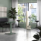 Moderní kancelářská scéna se zelenými stoly na notebooky MITTZON, černými stoličkami a šedou pohovkou s velkými okny a rostlinami.