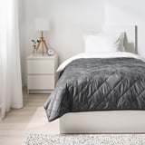 Moderní minimalistická ložnice: šedý přehoz na posteli mukplister na oddělené posteli, bílé polštáře, malý noční stolek s lampičkou, hodiny, rostlina. Podlaha ze světlého dřeva.