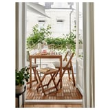 Malý balkon s dřevěnou jídelní soupravou, béžovými polštáři, rostlinami v květináčích a bílým výhledem na budovu.