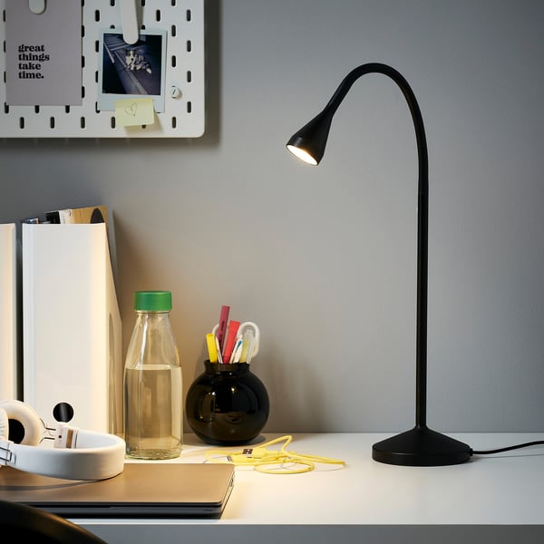 Moderní minimalistické uspořádání psacího stolu s černou lampou NÄVLINGE. Tenký, flexibilní design. Na stole jsou také sluchátka, láhev s vodou a kancelářské potřeby.