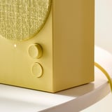 Detailní záběr elegantního, olivově zeleného bluetooth reproduktoru s látkovým krytem a kulatými ovládacími prvky, umístěného na stylovém zakřiveném povrchu.