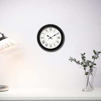 Moderní quartzové hodiny s černým rámem visí nad stolem s lampou a květinami.