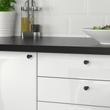 Moderní kuchyně s bílými skříňkami a černými deskami s černými knoflíky NYDALA, které umocňují styl skříňky. Na elegantní pracovní desce je salátová mísa.