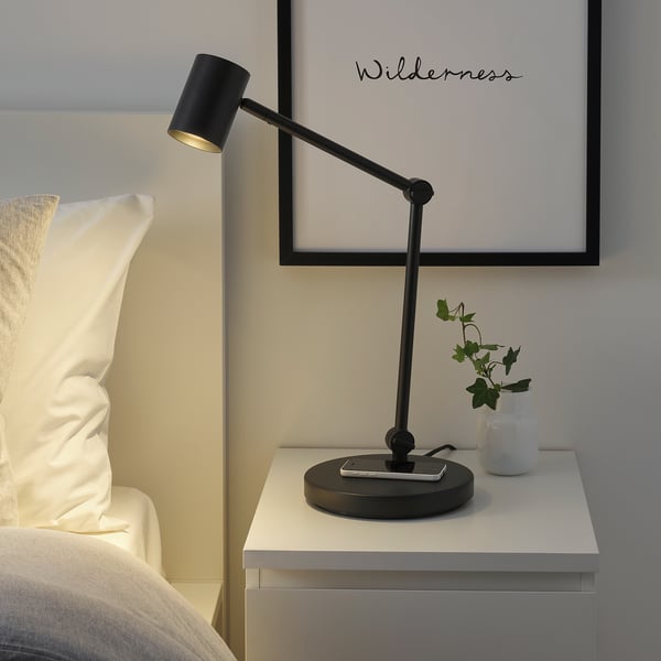 Moderní ložnice: černá lampa NYMÅNE, nastavitelná, bezdrátové nabíjení, na bílém nočním stolku vedle postele.