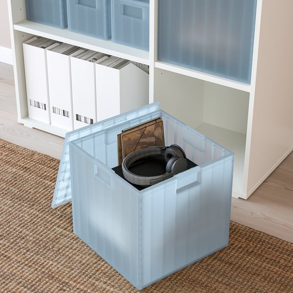 Průhledná plastová úložná schránka na podlaze, držící sluchátka a další předměty. Součást regálového systému s dalšími boxy a uspořádanými předměty.