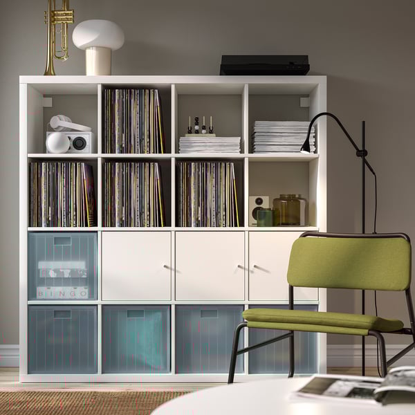 Moderní obývací pokoj s bílou knihovnou, vinylovými deskami a bytovými dekoracemi. Zelené křeslo a lampa v blízkosti, s uspořádaným úložným prostorem.