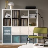 Moderní obývací pokoj s bílou knihovnou, vinylovými deskami a bytovými dekoracemi. Zelené křeslo a lampa v blízkosti, s uspořádaným úložným prostorem.