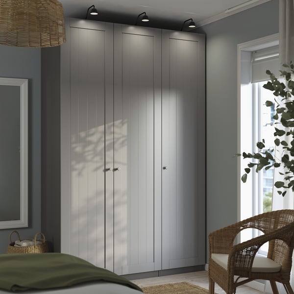 Moderní ložnice vysoká šedá šatní skříň PAX s elegantním designem, sladěným proutěným křeslem a zelenou přikrývkou na posteli.